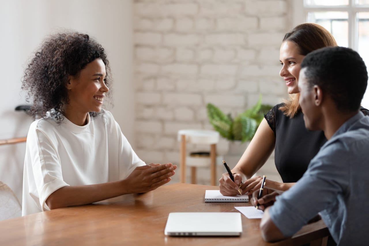 - A woman with curly hair and a white top is gesturing with her hands as she talks to the other two.
- A woman with brown hair and a black top is sitting next to a man with short black hair and a blue shirt.
- The man is holding a pen and writing in a notebook.
- There's a closed laptop and another notebook on the table.
- The background has a white brick wall, a plant, and a window letting in natural light.