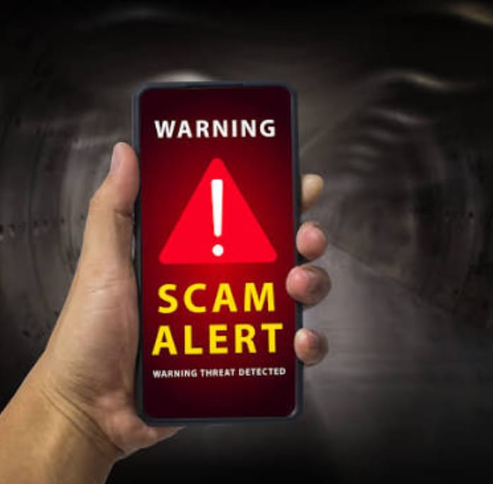 A hand's holding a smartphone with a big "SCAM ALERT" warning on the screen 🚨. The screen's got a red background with a red triangle and an exclamation mark in it. "WARNING" is written at the top in white, "SCAM ALERT" in yellow below the triangle, and "WARNING THREAT DETECTED" in small white text at the bottom. The background of the image is kinda dark and blurry