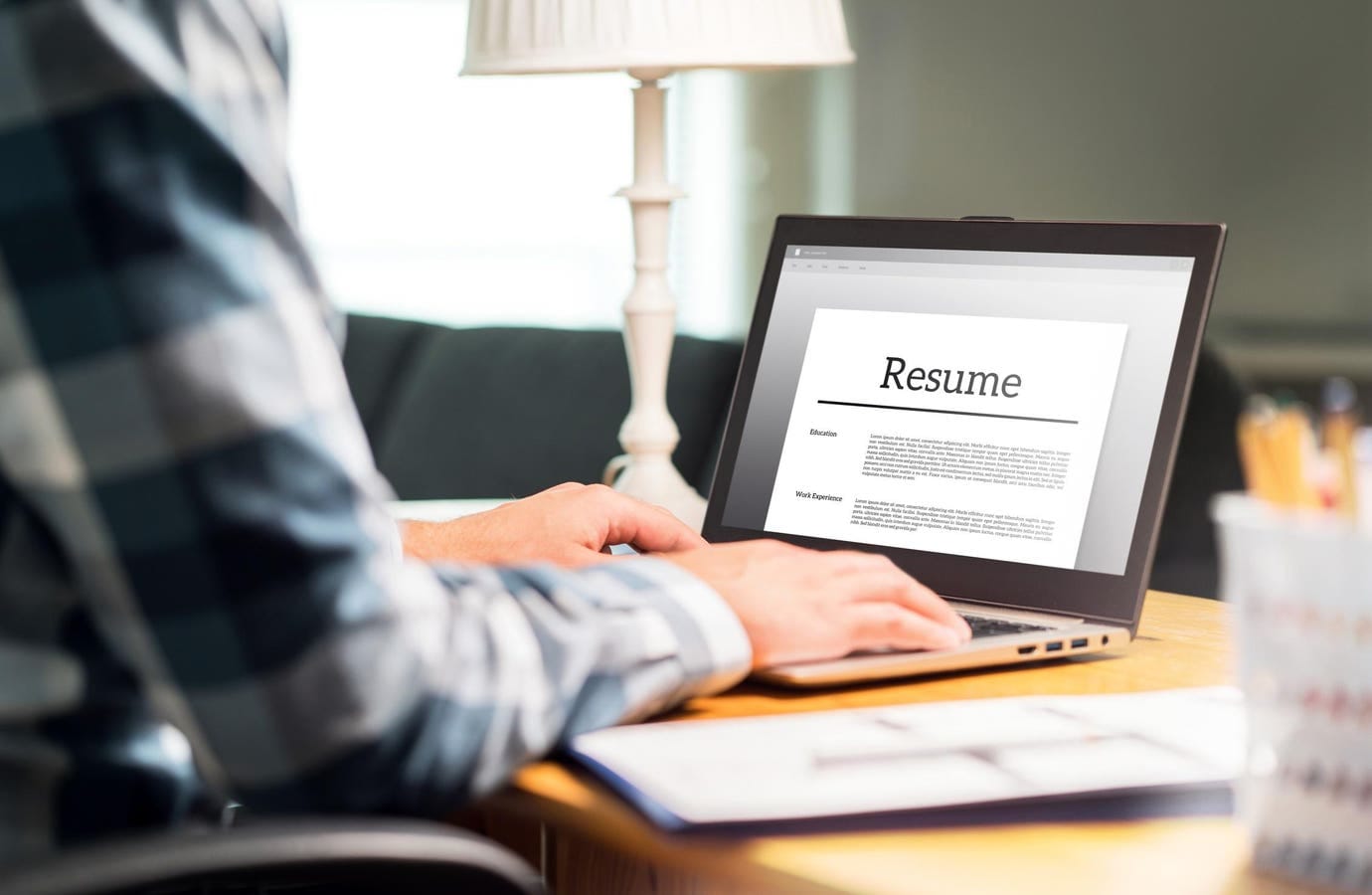 The image shows a person sitting at a desk with a laptop open, displaying a resume template with sections for "Education" and "Work Experience". There's a lamp in the background and some papers on the desk. 