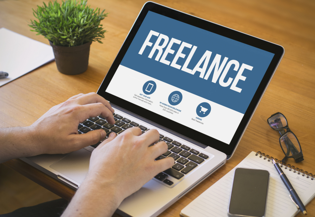 The image shows a person working on a laptop with the word "FREELANCE" prominently displayed on the screen.
- The laptop is on a wooden desk with a person's hands typing on the keyboard.
- The screen has a blue and white design with "FREELANCE" in large white letters.
- Below the title are three icons with text:
- "GET OUR APP" with a phone icon
- "WORK WORLDWIDE" with a globe icon
- "HIRING" with a shopping cart icon
- Other items on the desk include:
- A small potted plant (top left)
- A notebook and pen (bottom right)
- A smartphone (next to the notebook)
- A pair of glasses (next to the smartphone)