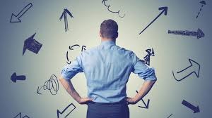 A guy's standin' with his back to us, hands on hips, lookin' at a wall full of crazy arrows pointin' every which way. The arrows are all diff shapes, sizes, and directions - like straight lines, curves, squiggles... wild stuff! The guy's wearin' a light blue shirt with rolled-up sleeves and dark pants. The wall's kinda greyish with a spotlight kinda effect on the center. Vibes are like he's thinkin', "Dude, which way do I go?"