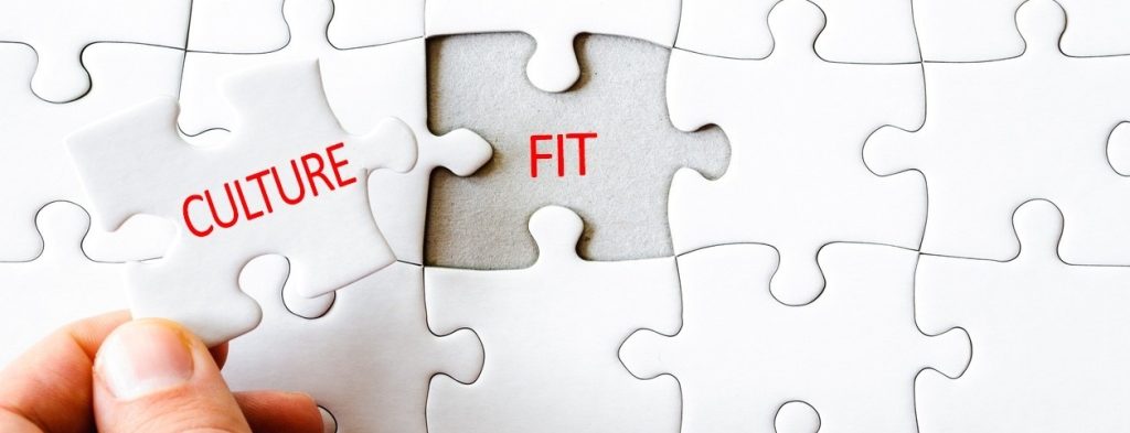 A close-up of a white jigsaw puzzle with one piece being placed into the puzzle. The piece has the word “CULTURE” in red, and the puzzle space it fits into has the word “FIT” in red, symbolizing the concept of cultural fit as the missing piece completing the organization.