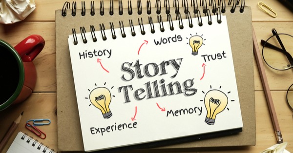A notebook placed on a wooden desk with the words “Story Telling” written in the center. Around it are hand-drawn light bulb icons and keywords like history, words, trust, memory, and experience, representing the key elements of storytelling. The image symbolizes creativity, learning, and the power of storytelling.