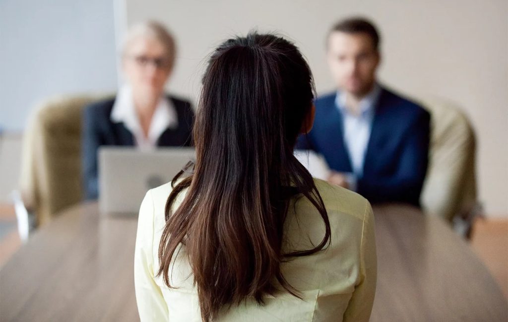A job candidate seen from behind, sitting at a table and facing two interviewers in a professional interview setting, representing a formal job interview or recruitment discussion.