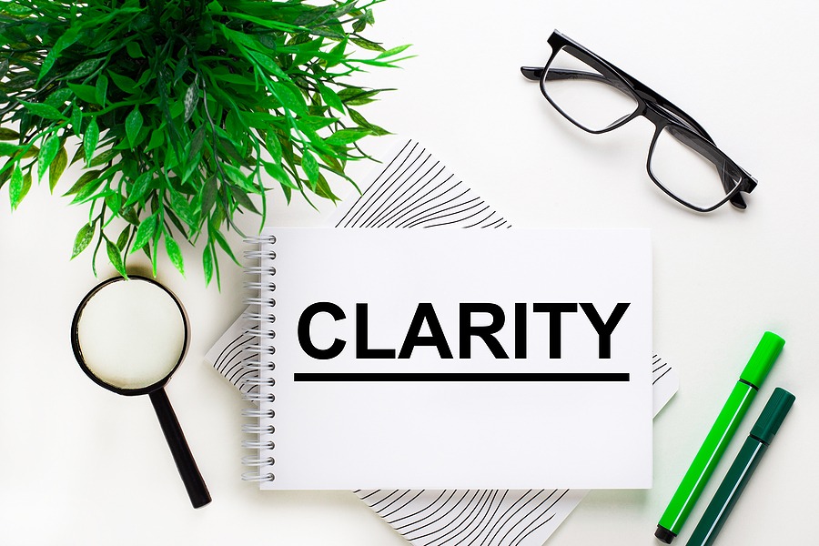 A notebook with the word “CLARITY” written in bold letters, placed on a clean desk with a magnifying glass, eyeglasses, green pens, papers, and a small plant, representing focus, organization, and clear thinking.