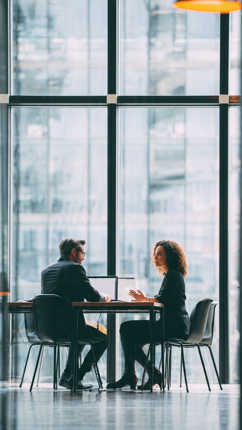 Questions to Ask Employees
A job candidate and a company employee sit across from each other in a modern office meeting room. They are engaged in a professional conversation at a conference table with a laptop and documents visible. Large windows provide natural light, creating a welcoming and professional workplace atmosphere.